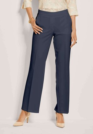 Vrouw in marineblauwe rechte broek, crèmekleurige textuur top, beige hoge hakken, gouden armband en donkere ring, staat met één hand in de zak.