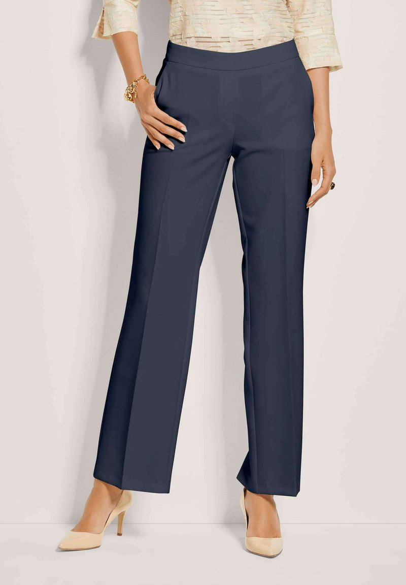 Vrouw in marineblauwe rechte broek, crèmekleurige textuur top, beige hoge hakken, gouden armband en donkere ring, staat met één hand in de zak.