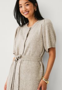Beige gebreide jurk met korte pofmouwen, ronde halslijn, knoopsluiting aan de voorkant en een stoffen riem die op de taille is gestrikt. Glad textuur, subtiel patroon.