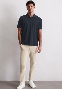 Dunkelgraues Poloshirt mit kurzen Ärmeln und Kragen, kombiniert mit hellbeigen Hosen und weißen Sneakers, steht vor einer strukturierten Wand.