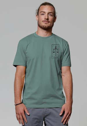 Green cotton t-shirt featuring a black graphic of scales and a sword, with a rectangular design on the left chest area. Short sleeves.