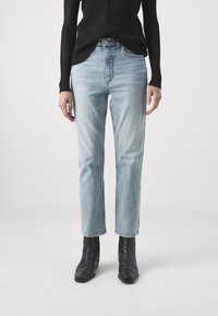 Osoba v těchto světlomodrých rovně střižených džínách, černém vroubkovaném svetru a černých kožených kotníkových botách, která stojí na jednoduchém bílém pozadí.