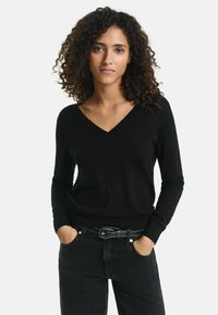 Schwarzer V-Ausschnitt-Pullover mit langen Ärmeln, aus einem weichen Material. Getragen mit hoch taillierten schwarzen Jeans und einem Gürtel in der Taille.