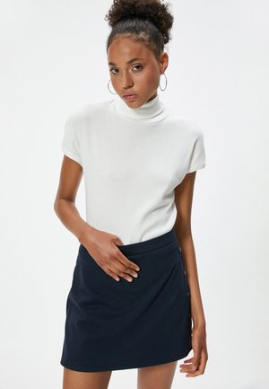 Top blanco de cuello vuelto de canalé combinado con una mini falda azul marino que presenta un detalle de botones en el lateral, textura suave y diseño ajustado.