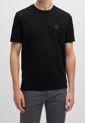 Schwarzes T-Shirt mit Rundhalsausschnitt aus weicher Baumwolle, mit einem kleinen schwarzen Logo-Patch auf der linken Brust, kurzen Ärmeln und regulärer Passform.