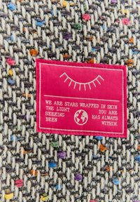 Tkanina s šedým tkaným vzorem proloženým barevnými skvrnami. Růžová textilní visačka zobrazuje bílý text a grafiku globu.