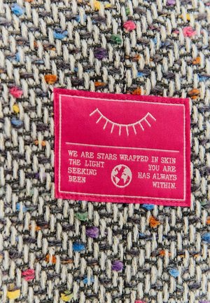 Tkanina z szarym wzorem tkanym, przerywanym kolorowymi plamkami. Różowa metka materiałowa z białym tekstem i grafiką kuli ziemskiej.