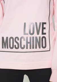 Light pink sweatshirt featuring "LOVE MOSCHINO" in bold black text with a dotted pattern, framed by a thin black border. Soft fabric.