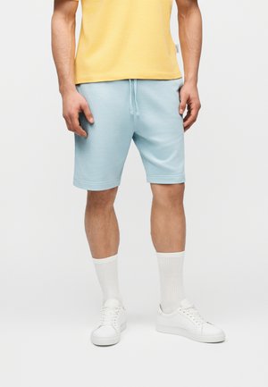 Άνδρας που φοράει ανοιχτό μπλε σορτς με κορδόνια, λευκά αθλητικά παπούτσια, λευκές κάλτσες και κίτρινη μπλούζα, στέκεται μπροστά σε μια απλή φόντο.