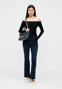 Woman wearing black off-shoulder top, dark blue flared jeans, white pointed shoes, and carrying a dark navy handbag on her shoulder.