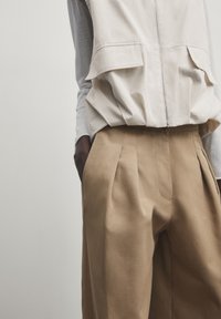 Gilet sans manches beige clair avec poches avant associé à un pantalon plissé beige. Tissu texturé, coupe légèrement ample, design minimaliste.