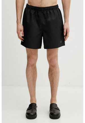 Uomo che indossa pantaloncini con vita elastica neri e scarpe slip-on nere, in piedi contro uno sfondo chiaro e semplice.