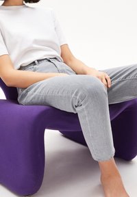 Kaareva violetti tuoli, jossa on pehmeä tekstuuri; henkilö pukeutunut istuvaan valkoiseen paitaan ja vaaleanharmaisiin farkkuihin, istuu jalat ristissä.