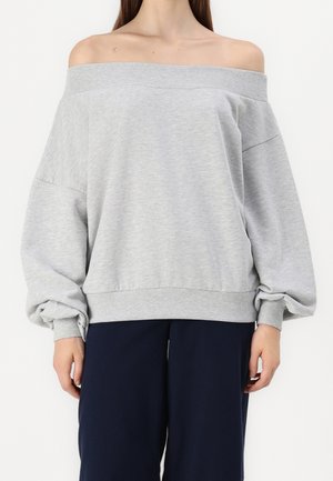 Kvinde iført en grå off-shoulder sweatshirt med lange ballonærmer og marineblå bukser med vide ben mod en ensfarvet baggrund.
