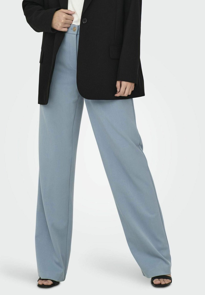 Pantalon ample bleu clair en tissu doux, associé à un blazer noir et un haut blanc. Le pantalon présente un bouton sur le devant et un design épuré.