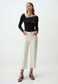 Zwart geribbeld longsleeve shirt met een asymmetrische halslijn en een decoratief detail. Witte broek met wijde pijpen, voorzakken en een knoopsluiting.