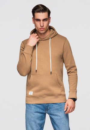 Młody mężczyzna z zaczesanymi do tyłu włosami, ubrany w beżową bluzę z białymi sznurkami i niebieskie dżinsy, poprawiający kołnierz bluzy lewą ręką.