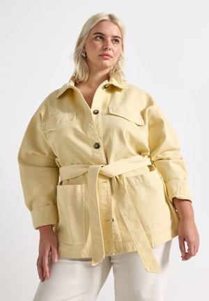 Kvinde med blondt hår iført en oversize lysegul jakke med bælte og store forlommer samt hvide bukser, der kigger opad mod venstre.