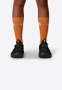 Zapatos de senderismo negros con suelas texturizadas, combinados con calcetas naranjas hasta la rodilla que cuentan con una marca blanca y un diseño de canalé vertical.
