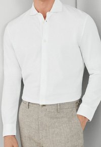 White button-up shirt with a narrow collar, long sleeves, and smooth texture, paired with light brown herringbone trousers featuring button accents.