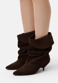 Stivali ankle in suede marrone con un design morbido, punta appuntita e tacco kitten basso. Texture morbida e risvolto piegato nella parte superiore.