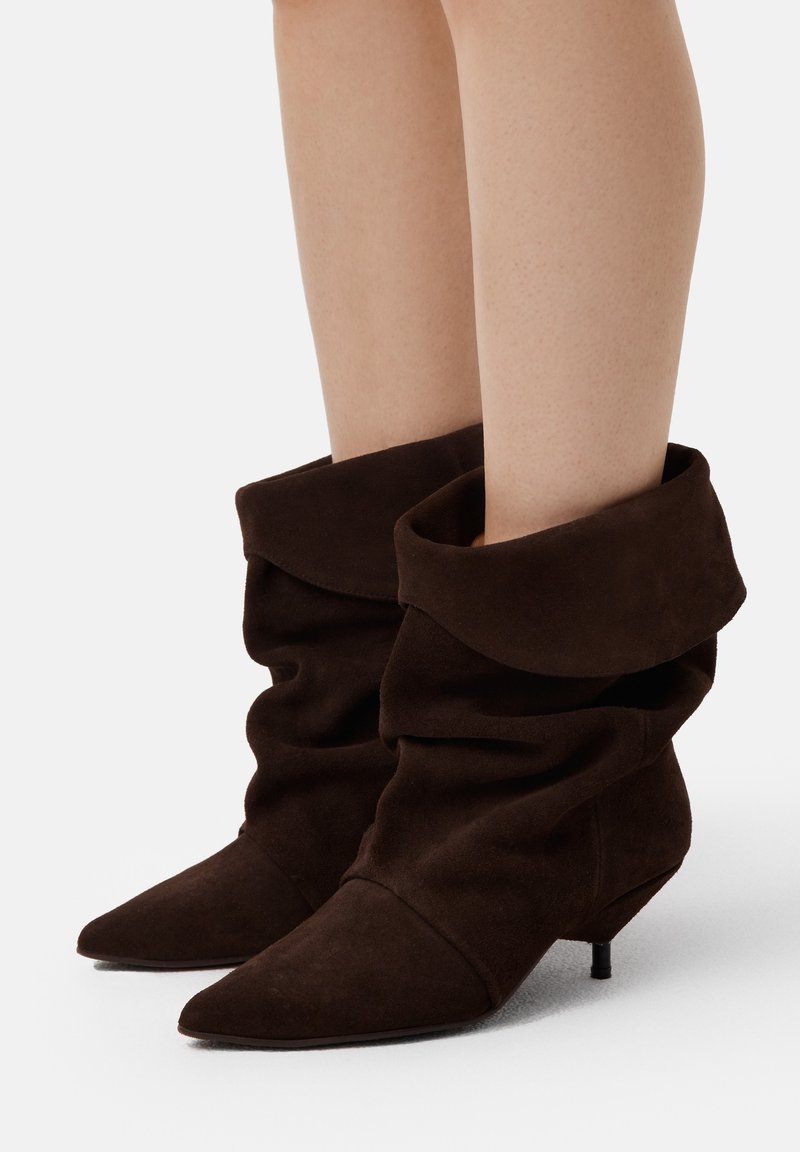 Stivali ankle in suede marrone con un design morbido, punta appuntita e tacco kitten basso. Texture morbida e risvolto piegato nella parte superiore.