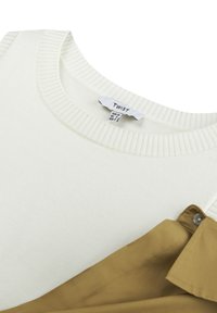Jasno beżowy sweter robiony na drutach z karczkiem ściągaczem, w połączeniu z brązową koszulą z kołnierzykiem z detalem guzika. Obie tkaniny są gładkie.