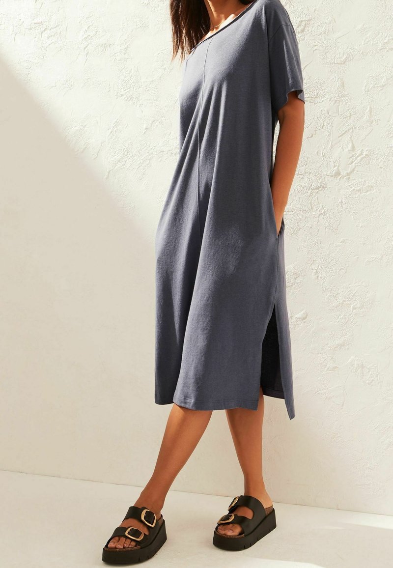 Robe bleu marine, longueur genou, avec des manches courtes, des poches latérales et une coupe décontractée ; portée avec des sandales à plateforme noires avec des boucles.