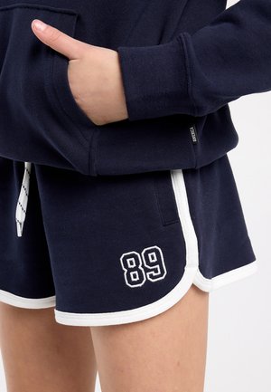 Persoon draagt marineblauwe korte broek met witte bies en nummer 89 op het been, hand gestoken in de zak van een bijpassende marineblauwe trui.