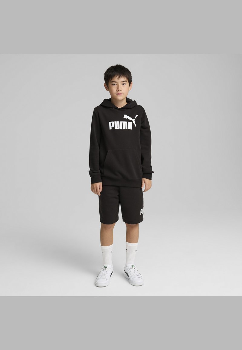 Crna dukserica s bijelim logotipom "PUMA", crne kratke hlače, bijele čarape i bijele tenisice. Ima kengursku džep i kapuljaču.