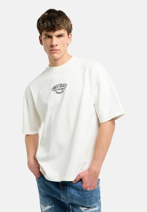 Ung mand iført hvid oversized T-shirt med sort tekstlogo og blå revnede jeans, stående med hænderne i lommerne mod en hvid baggrund.