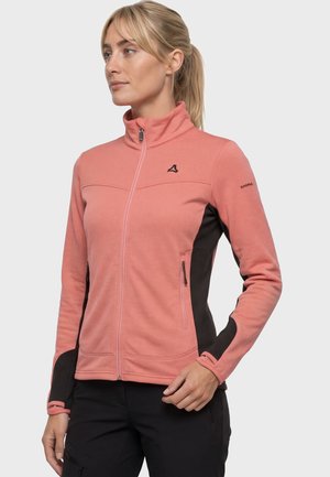 Frau trägt eine korallpinkfarbene Fleecejacke mit Reißverschluss, schwarzen Seitenpaneelen und schwarze Hose, steht mit einer Hand in der Hüfte.