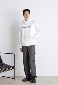 Witte hoodie met "Calvin Klein" logo, gedragen met donkergrijze cargobroek. Model staat tegen een effen achtergrond, zwarte schoenen en stoel zichtbaar.