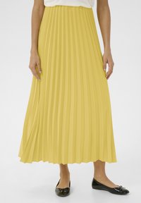 Gonna maxi plissettata gialla con superficie texturizzata, vita liscia e design fluido, abbinata a scarpe basse nere con un dettaglio a fiocco.