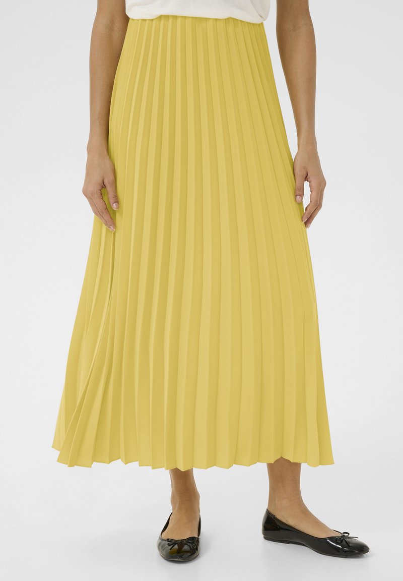 Gonna maxi plissettata gialla con superficie texturizzata, vita liscia e design fluido, abbinata a scarpe basse nere con un dettaglio a fiocco.