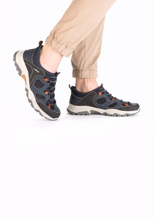 Chaussures de randonnée bleu marine avec des accents gris, semelles texturées, touches orange et lacets élastiques, assorties à un pantalon beige.