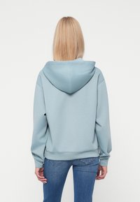Hanorac albastru deschis cu o glugă mare, textură moale, croială lejeră și manșete cu dungi. Asortat cu blugi, îmbinând un stil casual.