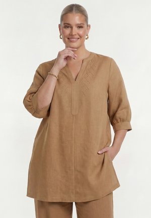 Kvinde med lys hud og blondt hår, iført guldbøjleøreringe, et armbånd og en løs tan tunika og bukser, smilende med hånden nær ansigtet.