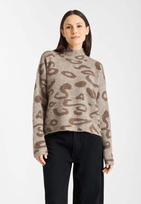 Gebreide trui in lichtbruin met donkere abstracte patronen, voorzien van een hoge halslijn en een cropped ontwerp, gecombineerd met zwarte pants.