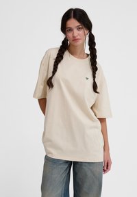 Beige oversized T-shirt lavet af bomuld, med korte ærmer, rund hals og et lille grønt broderet logo på brystet.