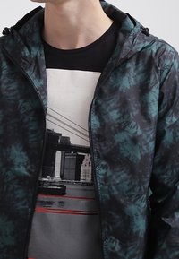 Wodoodporna kurtka z zielono-czarnym wzorem tie-dye, zapinana na zamek błyskawiczny, z regulowanym kapturem, noszona na t-shircie z grafiką.