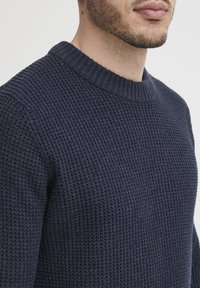 Sötétkék kötött pulóver texturált mintázattal, bordás kerek nyakkal és laza szabással, amely a melegség és a kényelem hangsúlyozására szolgál.