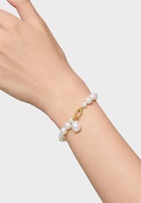 Bracciale in perle bianche con componenti in oro; presenta perle rotonde e a forma di goccia. Chiusura in oro, indossato su una mano.