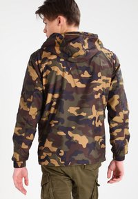Chaqueta de camuflaje con capucha, que presenta tonos de verde, marrón y amarillo. El material parece suave con puños elásticos, diseñada para la durabilidad.