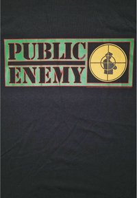 Černé bavlněné tričko s odvážnými blokovými písmeny "PUBLIC ENEMY" v červené a zelené barvě, s kruhovou grafikou postavy uvnitř zaměřovacího kříže.