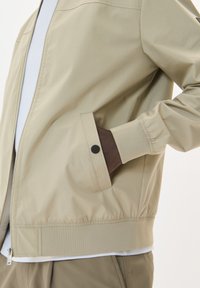 Chaqueta bomber beige con cierre frontal, puños elásticos y un bolsillo con un botón a presión negro. Presenta una textura suave y ligeramente brillante.