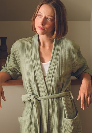 Frau mit kurzen braunen Haaren, die einen hellgrünen, strukturieren Bademantel trägt, der in der Taille gebunden ist, und sich drinnen auf eine Holzoberfläche stützt.