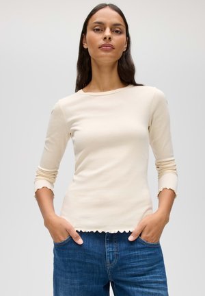 Donna con capelli lunghi e scuri che indossa una camicia crema a maniche lunghe con bordi arruffati e jeans blu, in piedi con le mani nelle tasche.