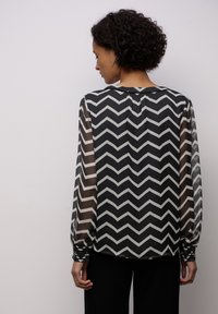 Blůza s černobílým Chevron vzorem, poloprůhlednými rukávy a nařasenými manžetami, s kulatým výstřihem a volným střihem.