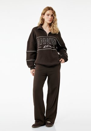 Joven mujer con cabello rubio ondulado que lleva una sudadera marrón oversize con el texto "TANNOV" y pantalones anchos a juego, de pie sobre fondo blanco.